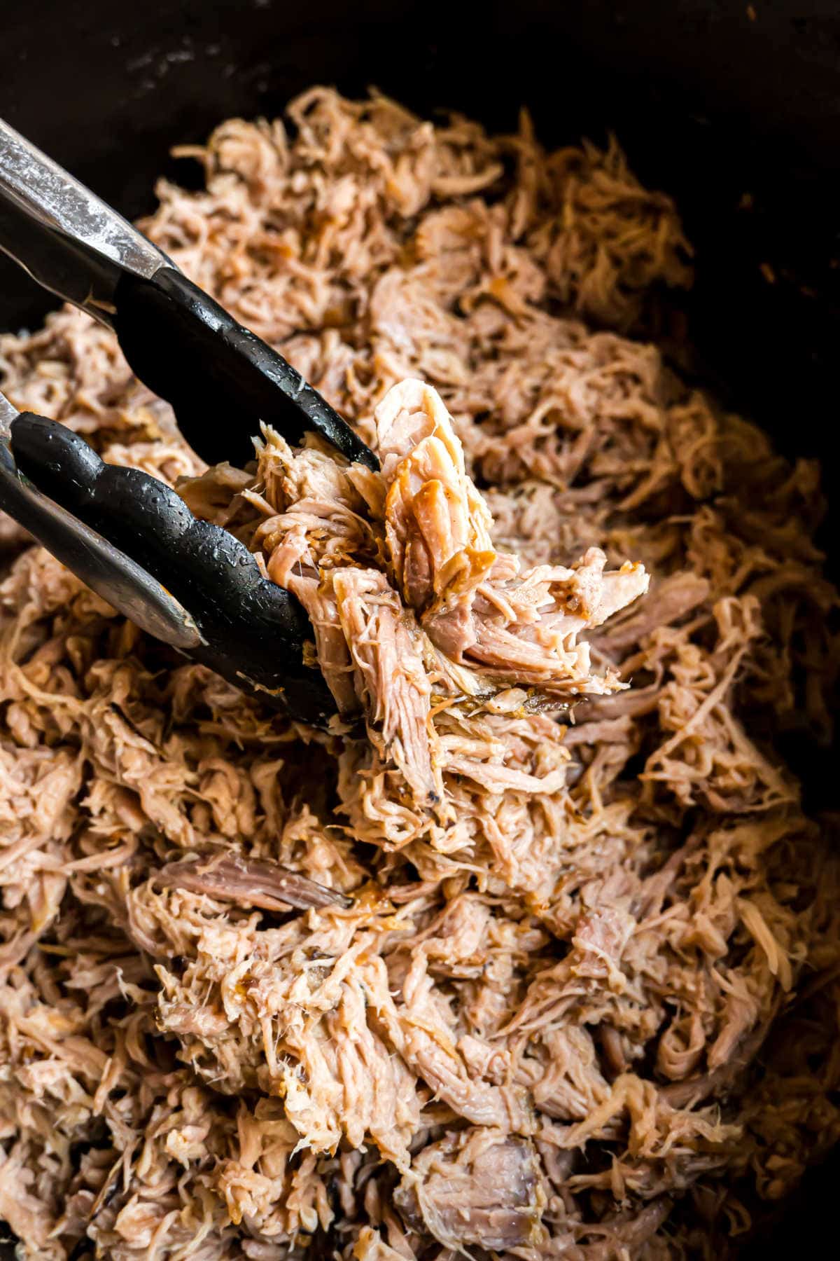 Shredded pork in a crockpot with a pair of tongs.