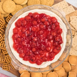 A wooden board boasts a glass dish brimming with cherry cheesecake dip, flanked by an assortment of cookies, pretzels, and graham crackers.