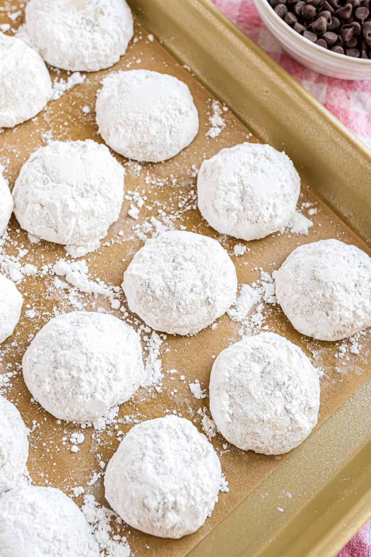 Chocolate chip snowball cookies, dusted in powdered sugar, are artfully arranged on a parchment-lined baking sheet. A bowl of chocolate chips peeks from the corner, adding a tempting hint of indulgence.