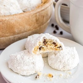 Chocolate chip snowball cookies dusted with powdered sugar, one broken to reveal the rich chocolate chips inside, rest on a white plate with a wooden bowl and cup in the background.