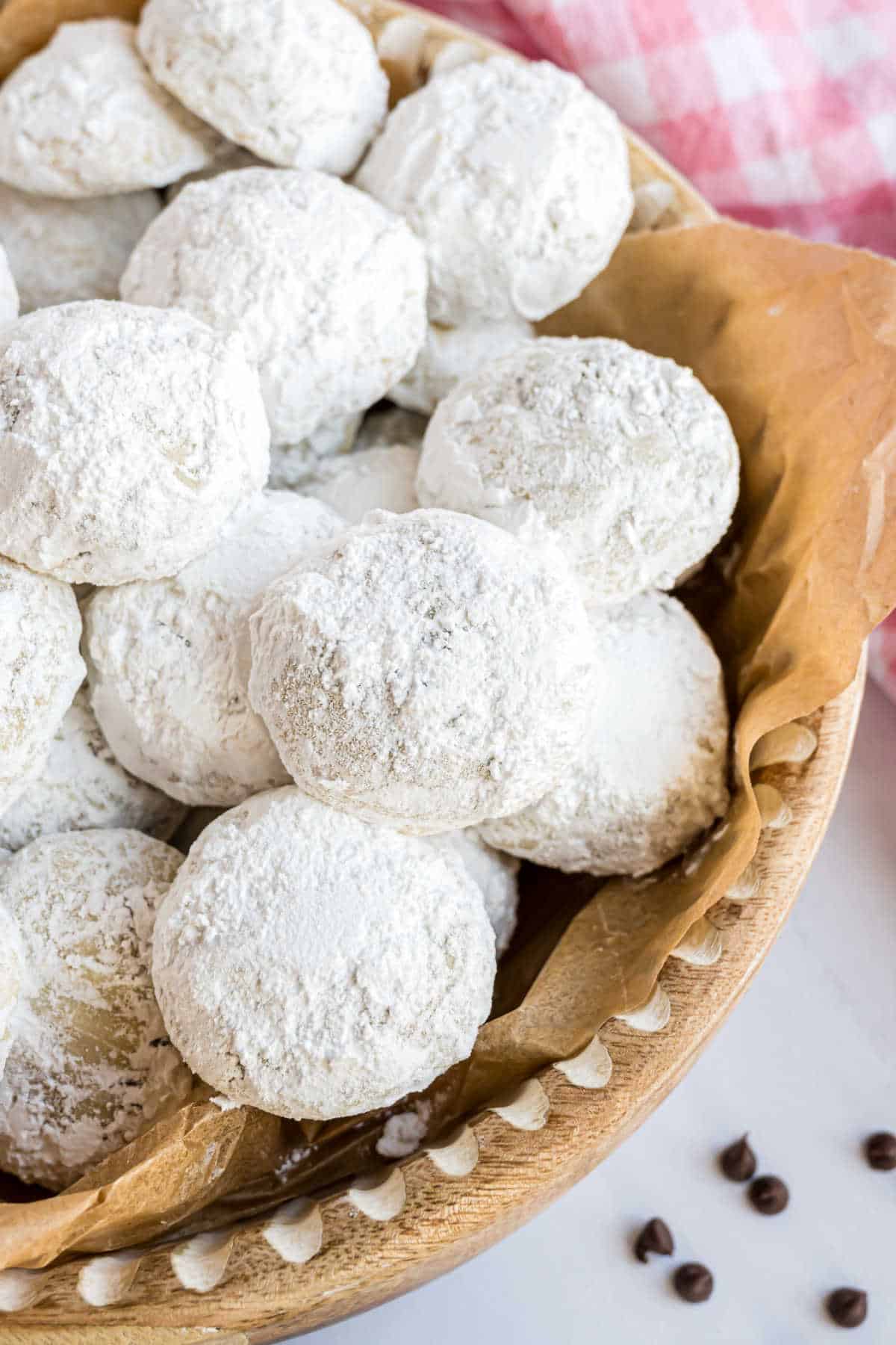 A wooden bowl filled with chocolate chip snowball cookies dusted in powdered sugar sits on brown paper, while a pink and white checkered cloth adds a charming backdrop.