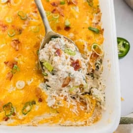 A jalapeño popper chicken casserole with melted cheese, bacon, and green onions is scooped with a spoon in a white dish.