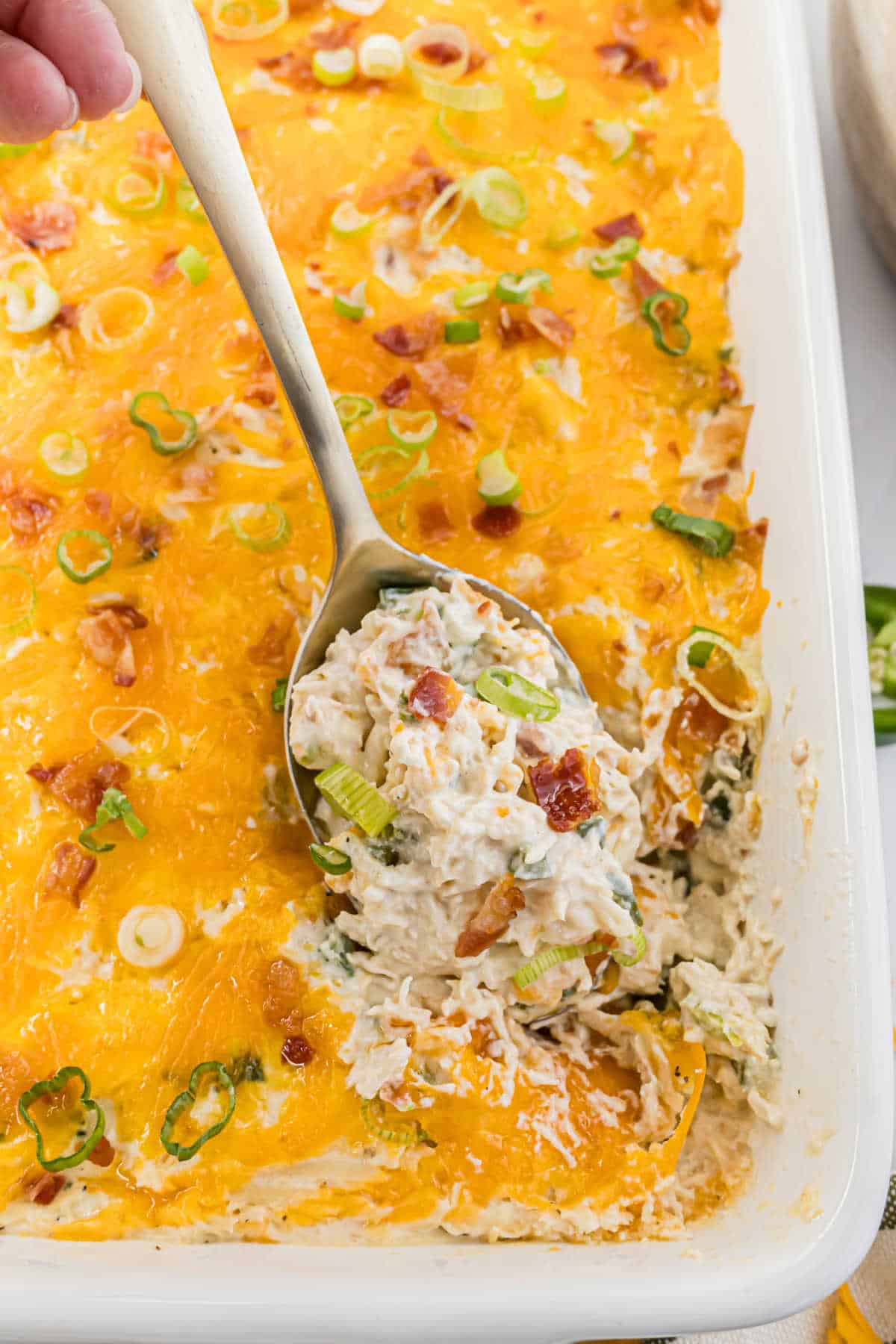 A spoon scoops the cheesy jalapeño popper chicken casserole, topped perfectly with crispy bacon and fresh green onions.