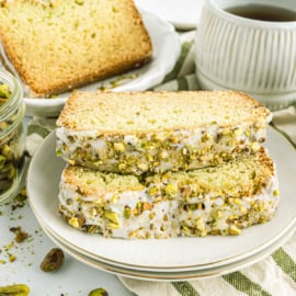 Two slices of pistachio cake with rich icing sit enticingly on a plate, reminiscent of freshly baked pistachio bread, accompanied by a steaming cup of coffee and scattered pistachios.