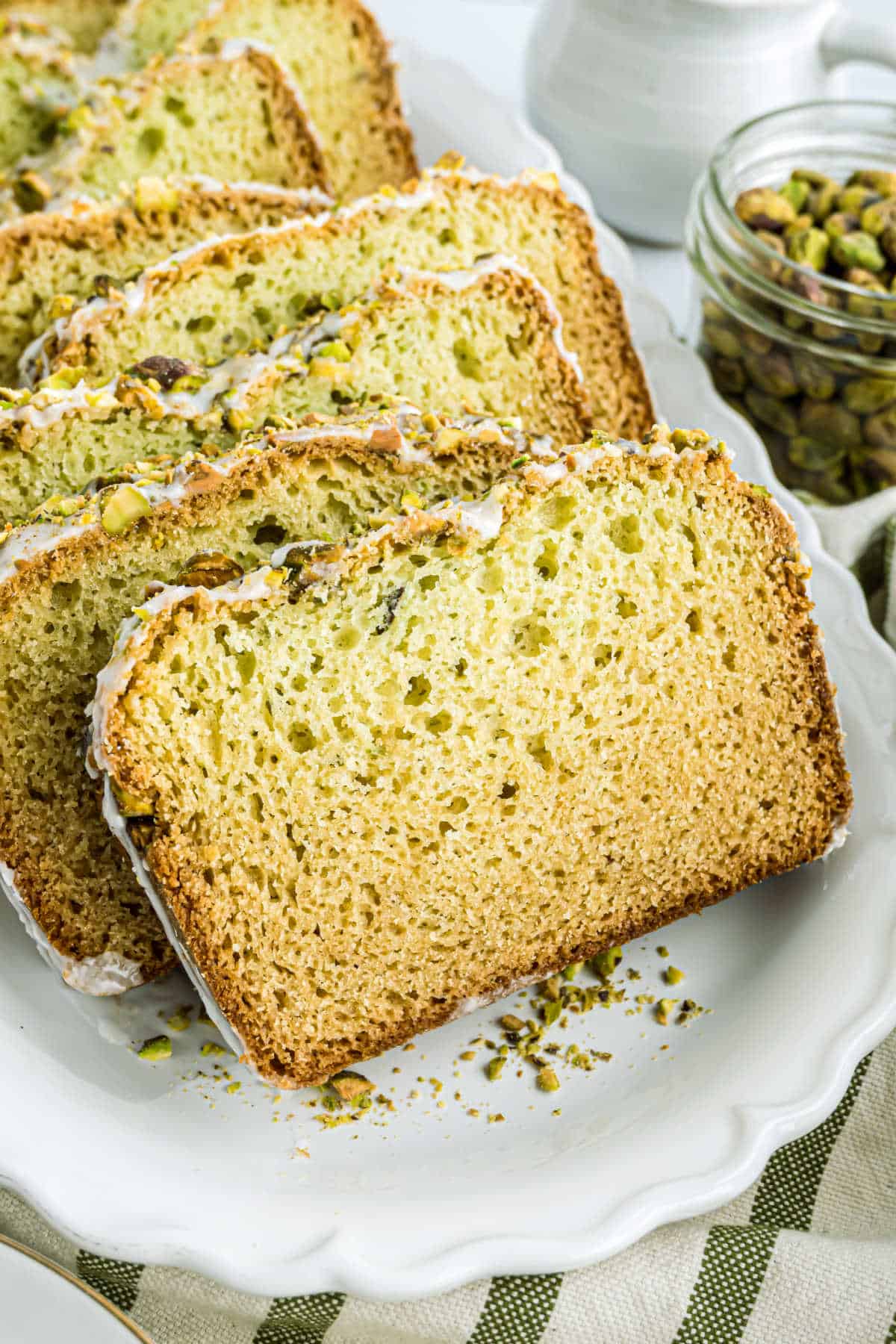 Pistachio bread slices with a drizzle of icing rest enticingly on a white plate, beside a jar brimming with vibrant pistachios.