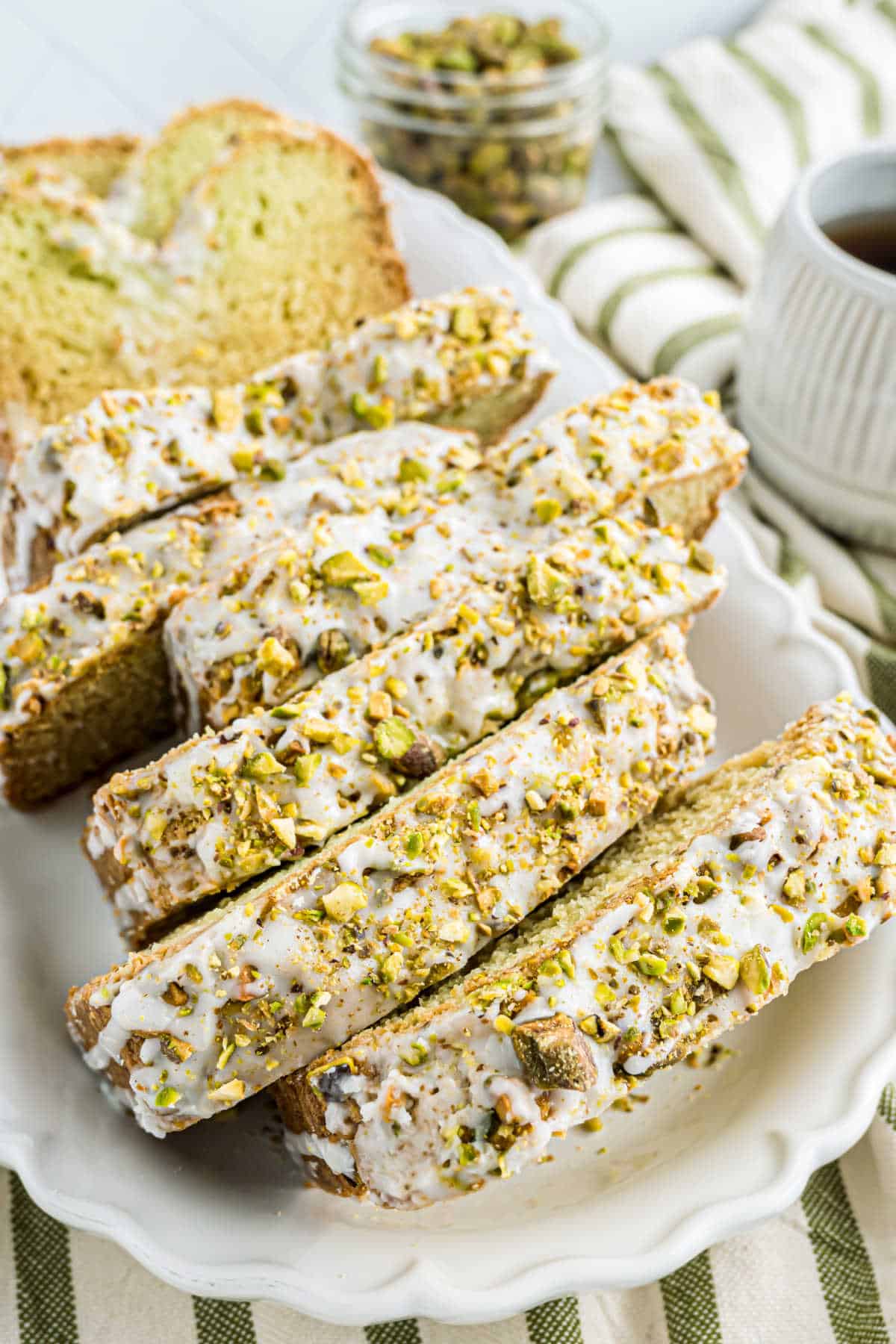 Sliced pistachio bread with sweet icing rests gracefully on a white plate, accompanied by a steaming cup of coffee and a jar overflowing with fresh pistachios.