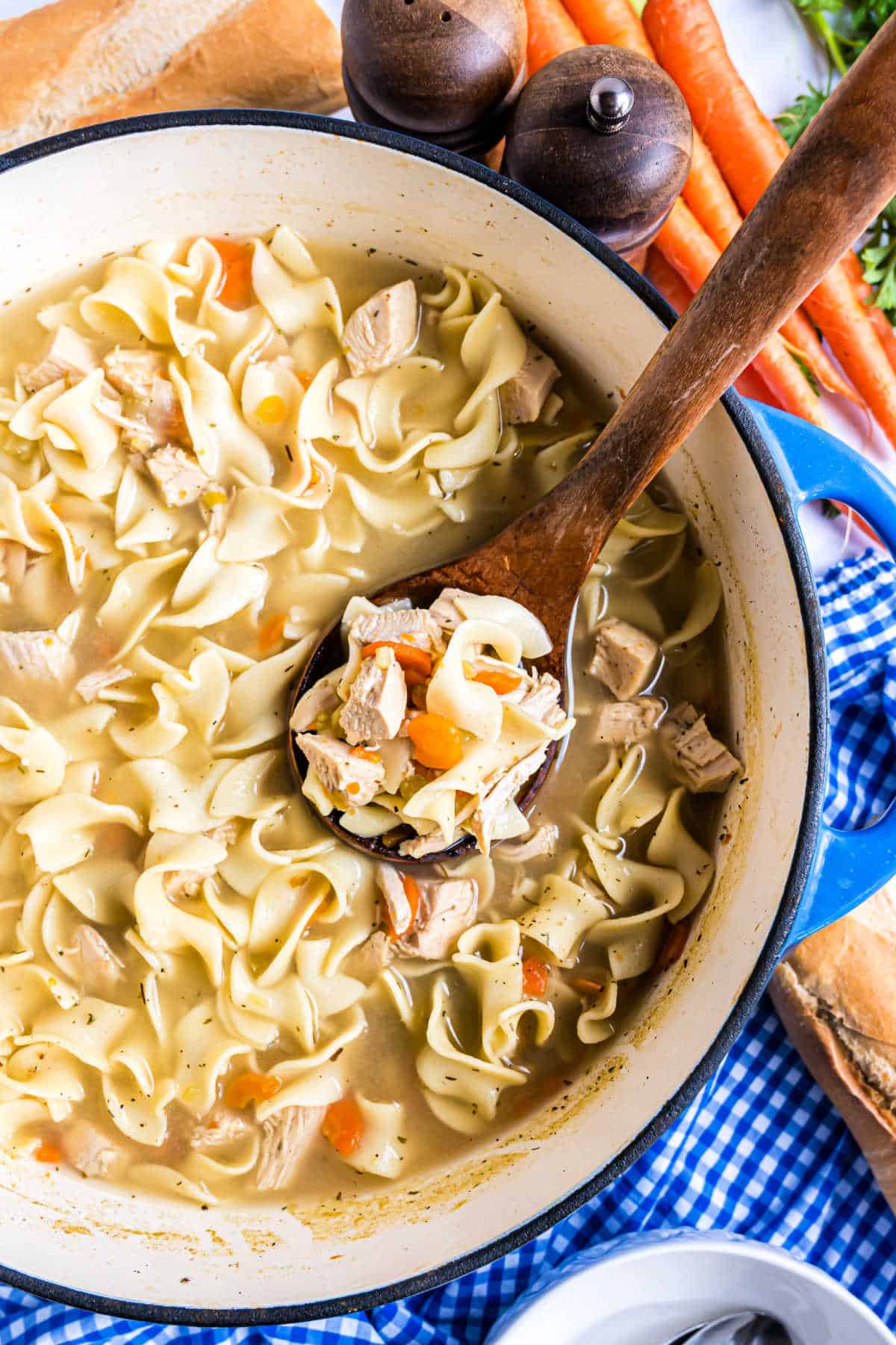 A comforting pot of chicken noodle soup, brimming with tender carrots and a wooden spoon resting on a blue checkered cloth, promises warmth and nourishment.