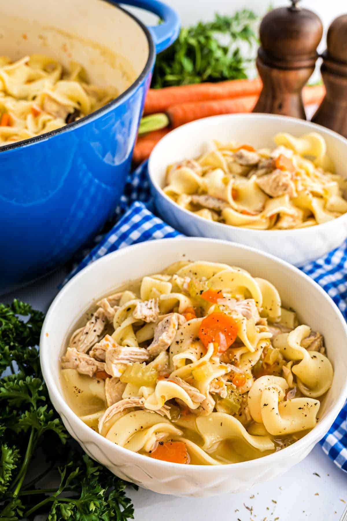 Two bowls of comforting chicken noodle soup with vegetables sit next to a blue pot, flanked by pepper grinders.