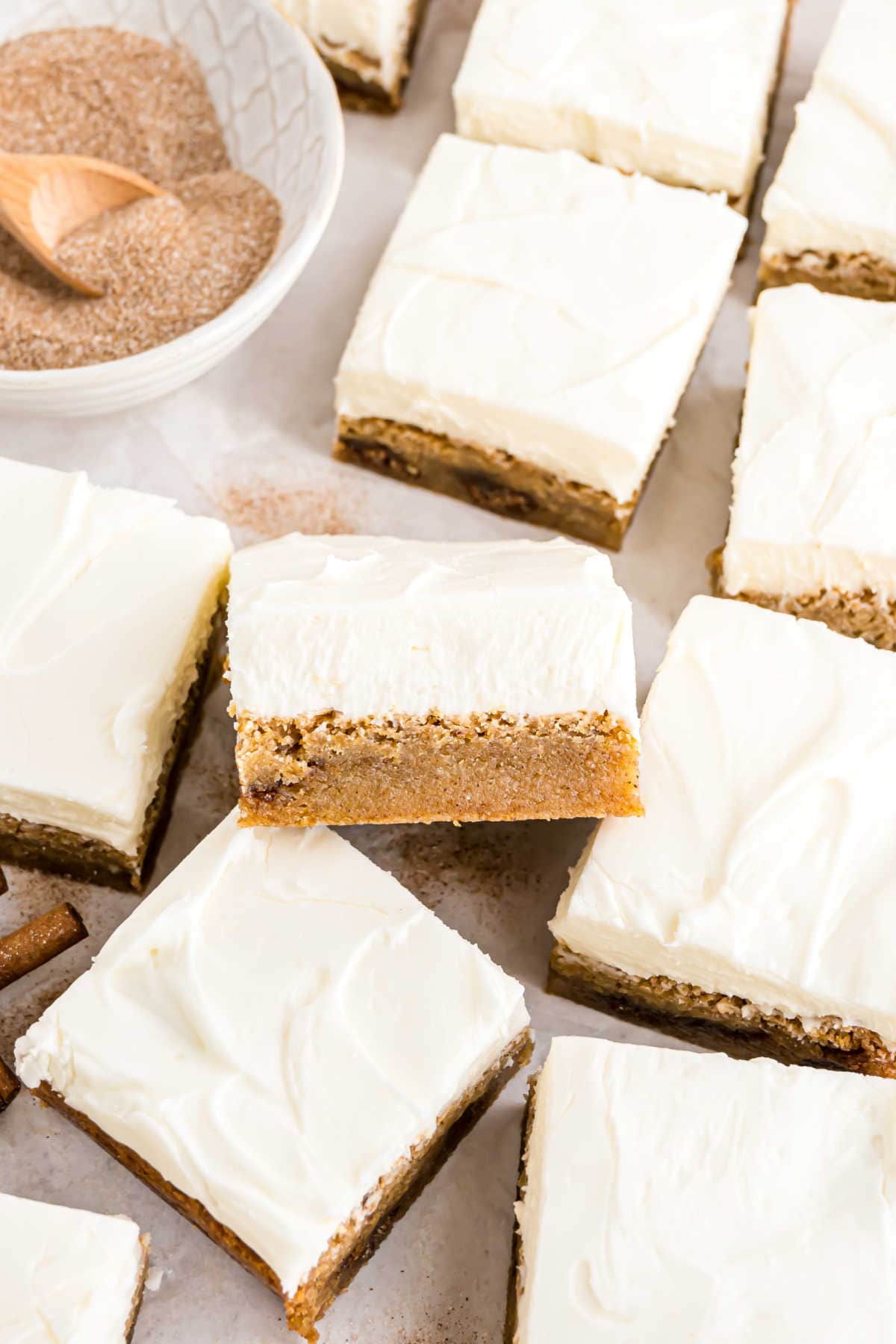 Frosted squares of pumpkin bars rest on a table, with a bowl of cinnamon sugar in the background offering a hint of sweetness, reminiscent of cinnamon roll blondies freshly baked.