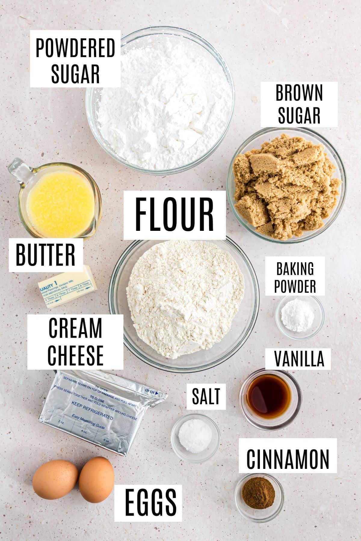 Baking ingredients for cinnamon roll blondies spill across the table: flour, brown sugar, powdered sugar, butter, eggs, cream cheese. A hint of fragrant spices and vanilla promise deliciousness.