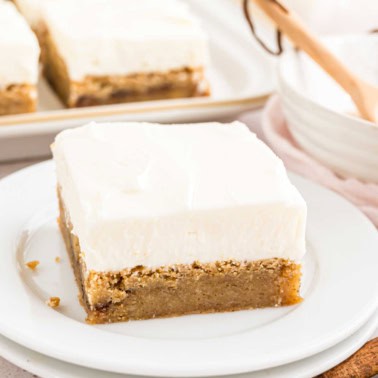 A square piece of frosted cinnamon roll blondie rests on a white plate, topped with a creamy layer that hints at a swirl of spice.