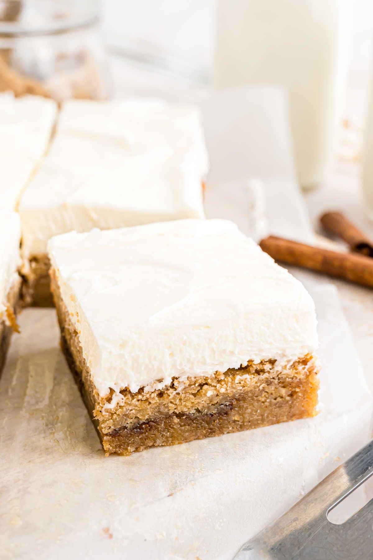 A slice of frosted cake on parchment paper with a creamy topping, surrounded by the charm of cinnamon roll blondies and baking utensils.